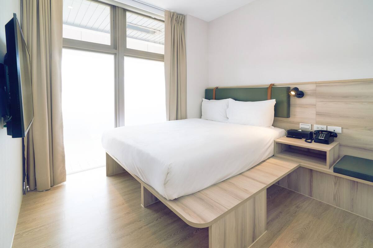 A modern hotel room with a bed, bedside table, and a view of the outdoors.