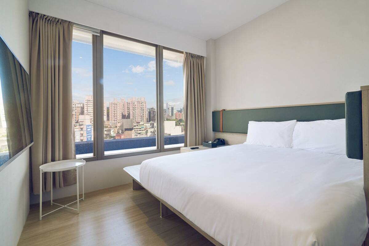A hotel room with a large bed, a small table, a nightstand, and a view of the city through large windows.