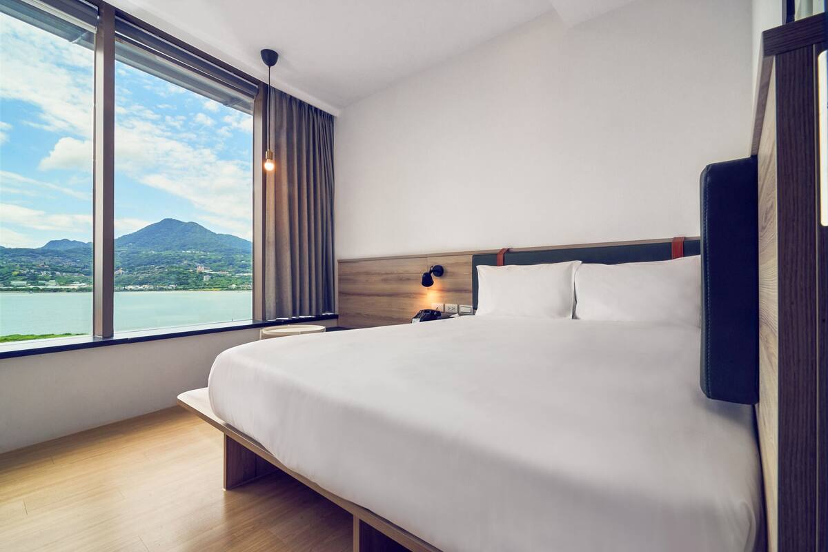 A modern hotel room with a large bed, a nightstand, a lamp, and a view of a lake and mountains.
