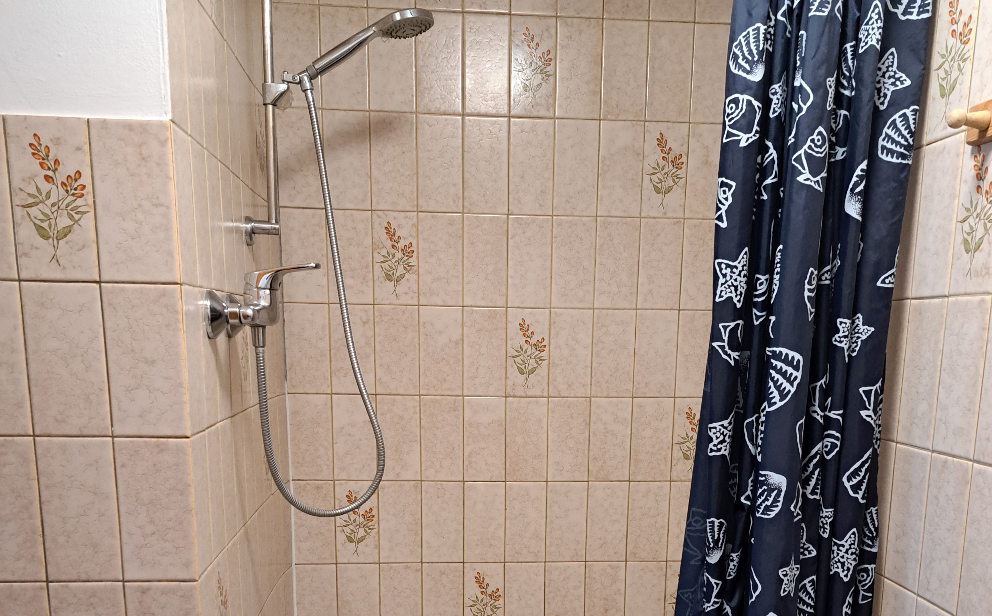 A shower area with beige tiles and floral patterns, a showerhead, and a dark blue shower curtain with fish designs.