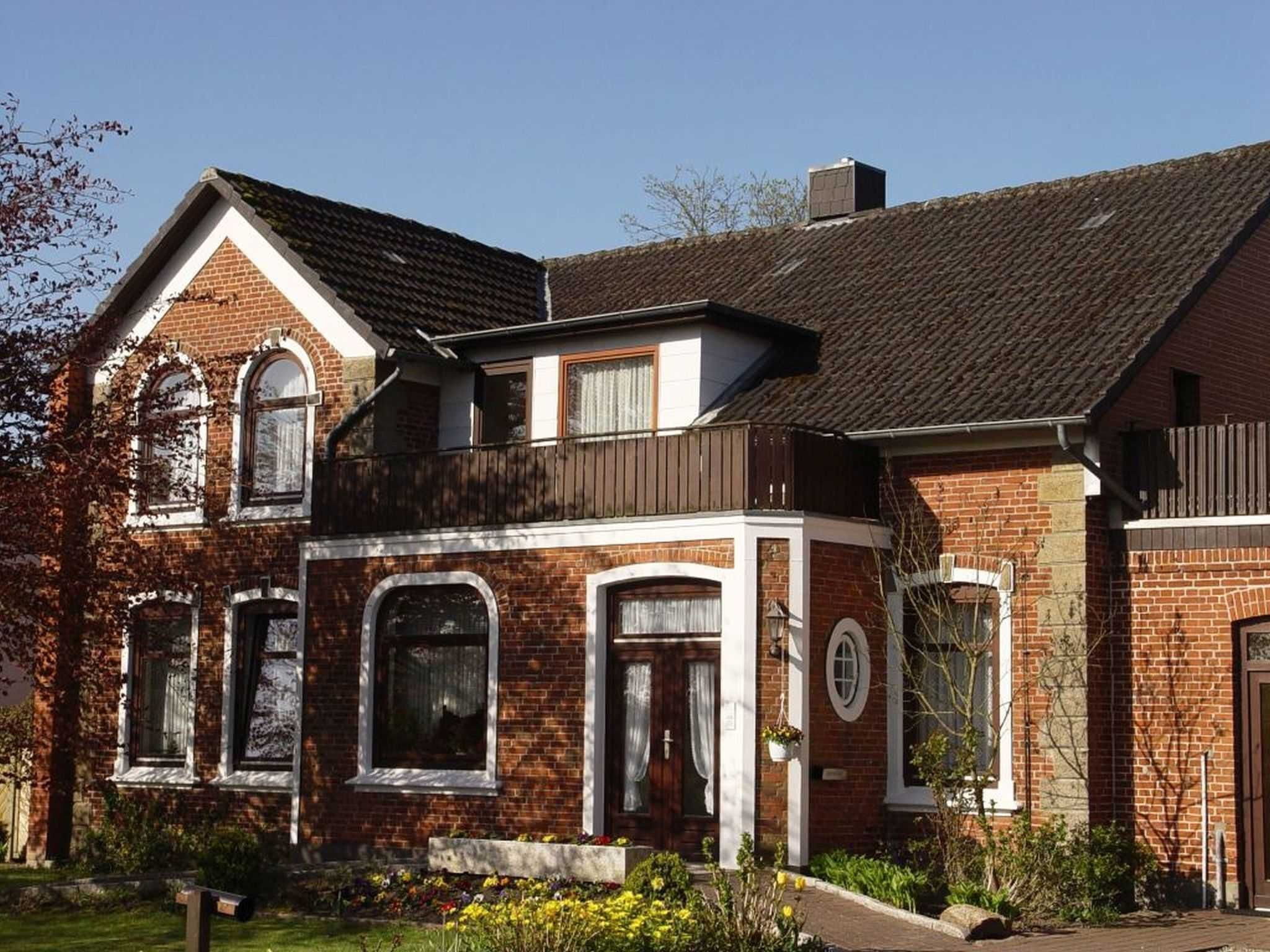 Une maison à deux étages, construite en briques, avec un toit à pignon, de grandes fenêtres et un porche à l’avant agrémenté d’un parterre de fleurs.