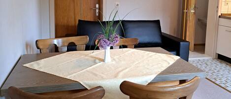 Uma sala de jantar com uma mesa quadrada, quatro cadeiras de madeira, um sofá preto e um vaso com flores roxas.