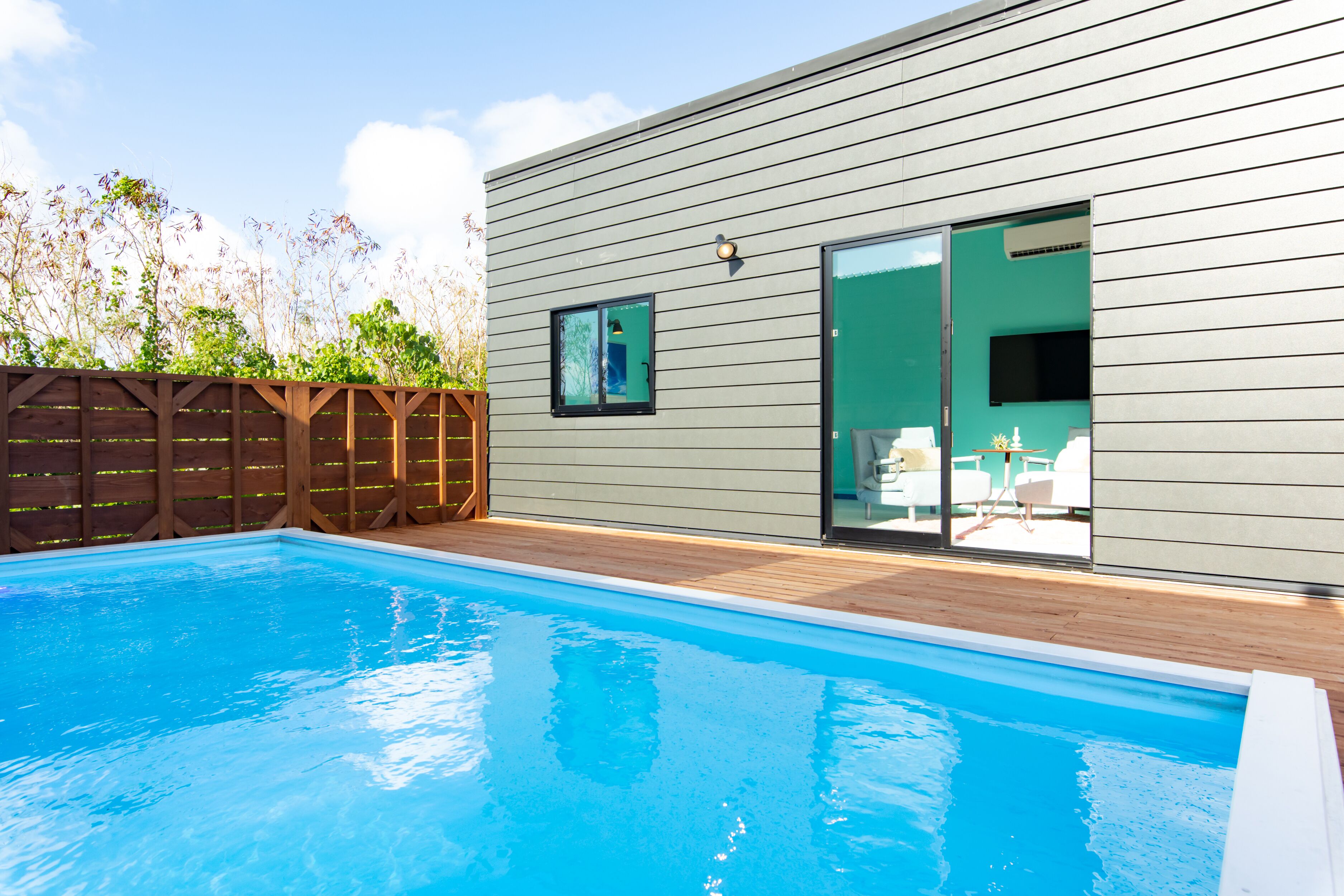 A modern outdoor pool area with a wooden deck and a sliding glass door leading to a patio with outdoor furniture.