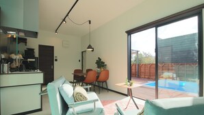 Une cuisine moderne avec un coin repas, une grande porte-fenêtre coulissante donnant sur un jardin avec piscine, et un coin salon confortable agrémenté d’un tapis rose.