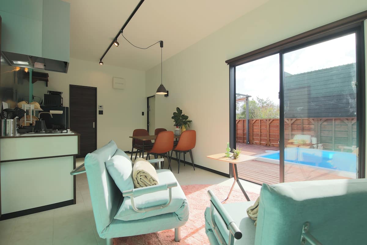 Una cocina moderna con comedor, un amplio ventanal corredero a un patio trasero con piscina y una zona de estar acogedora con alfombra rosa.