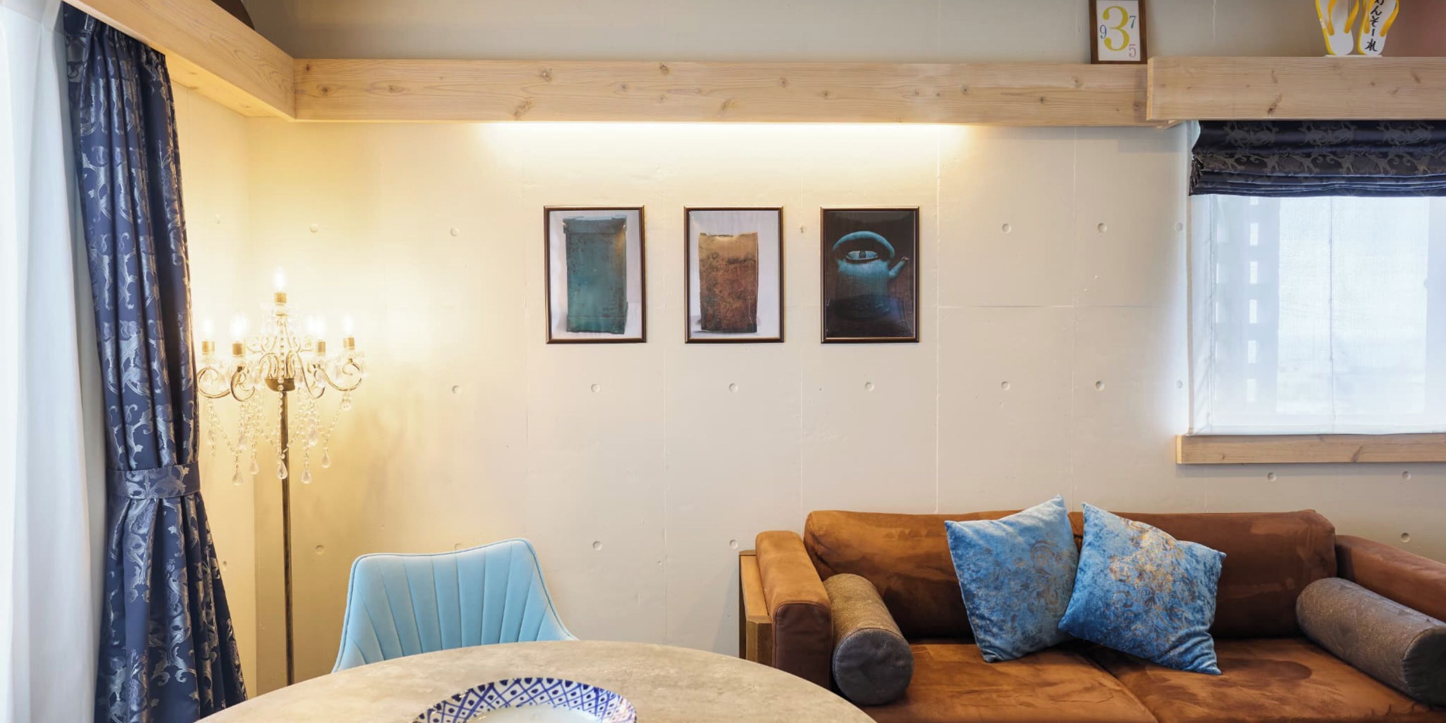 A room with a wooden ceiling, a brown sofa with patterned cushions, a blue chair, a chandelier, and three framed artworks on the wall.