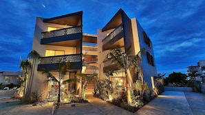 A modern multi-story building with balconies, surrounded by tropical plants and a well-lit entrance. - Yomitan Condhotel ND (Yomitan)