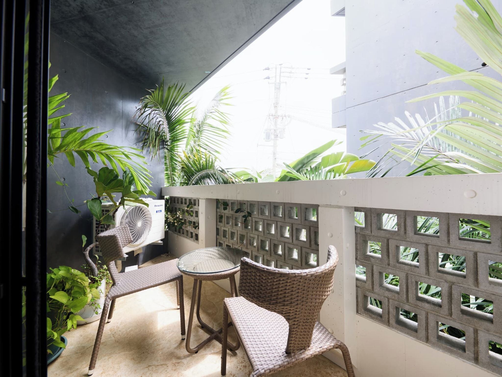 Un balcón con mobiliario de mimbre, plantas en macetas y una pared de concreto con diseño de cuadrícula.