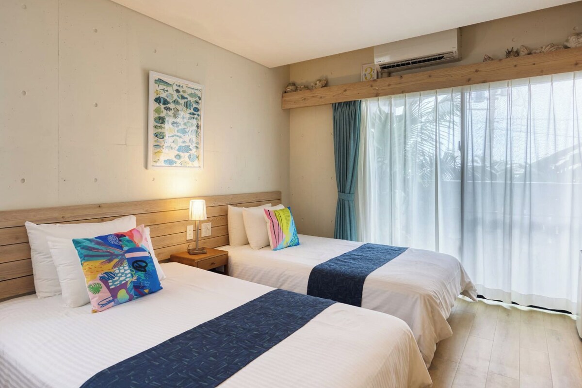 A hotel room with two beds, a wooden headboard, a wall-mounted air conditioner, a window with curtains, and a framed picture on the wall.
