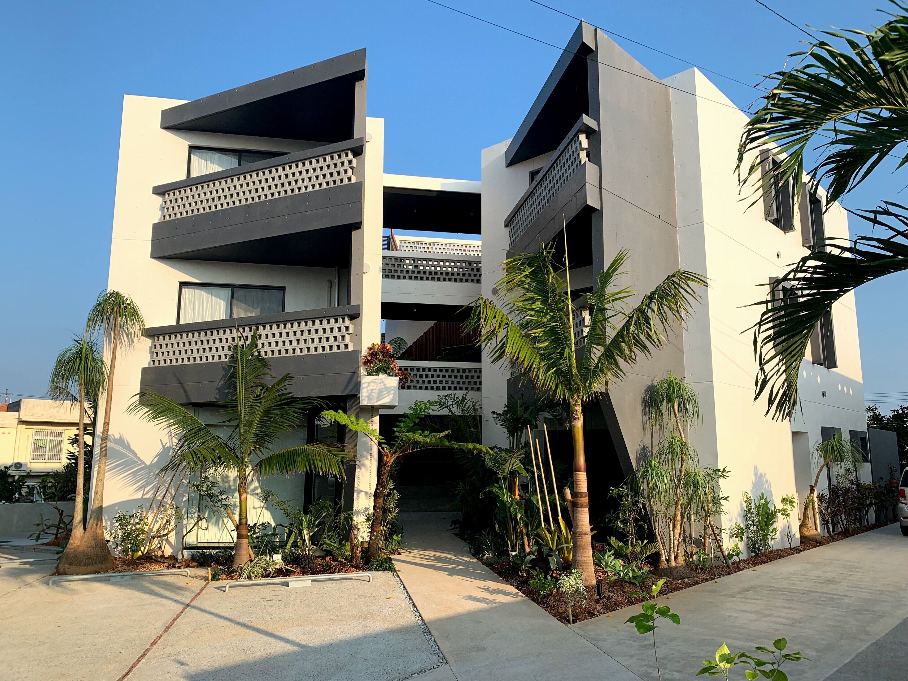 A modern multi-story building with balconies, surrounded by palm trees and a concrete pathway.