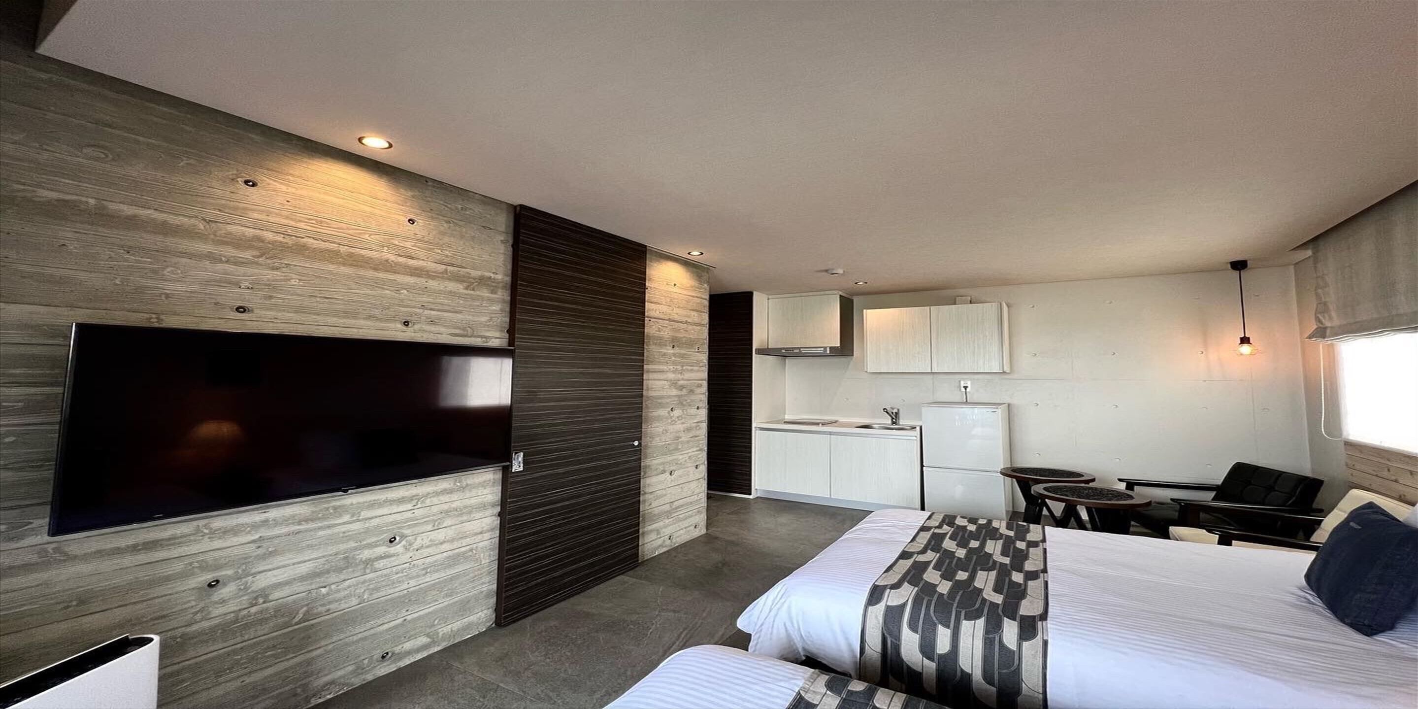 A modern hotel room with a large flat-screen TV, a bed with patterned bedding, a small kitchenette, and a seating area with a table and chairs.