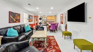 A modern living room with a black leather sofa, a wooden coffee table, and a vibrant red rug. There is a dining area with purple chairs and a kitchen visible in the background.