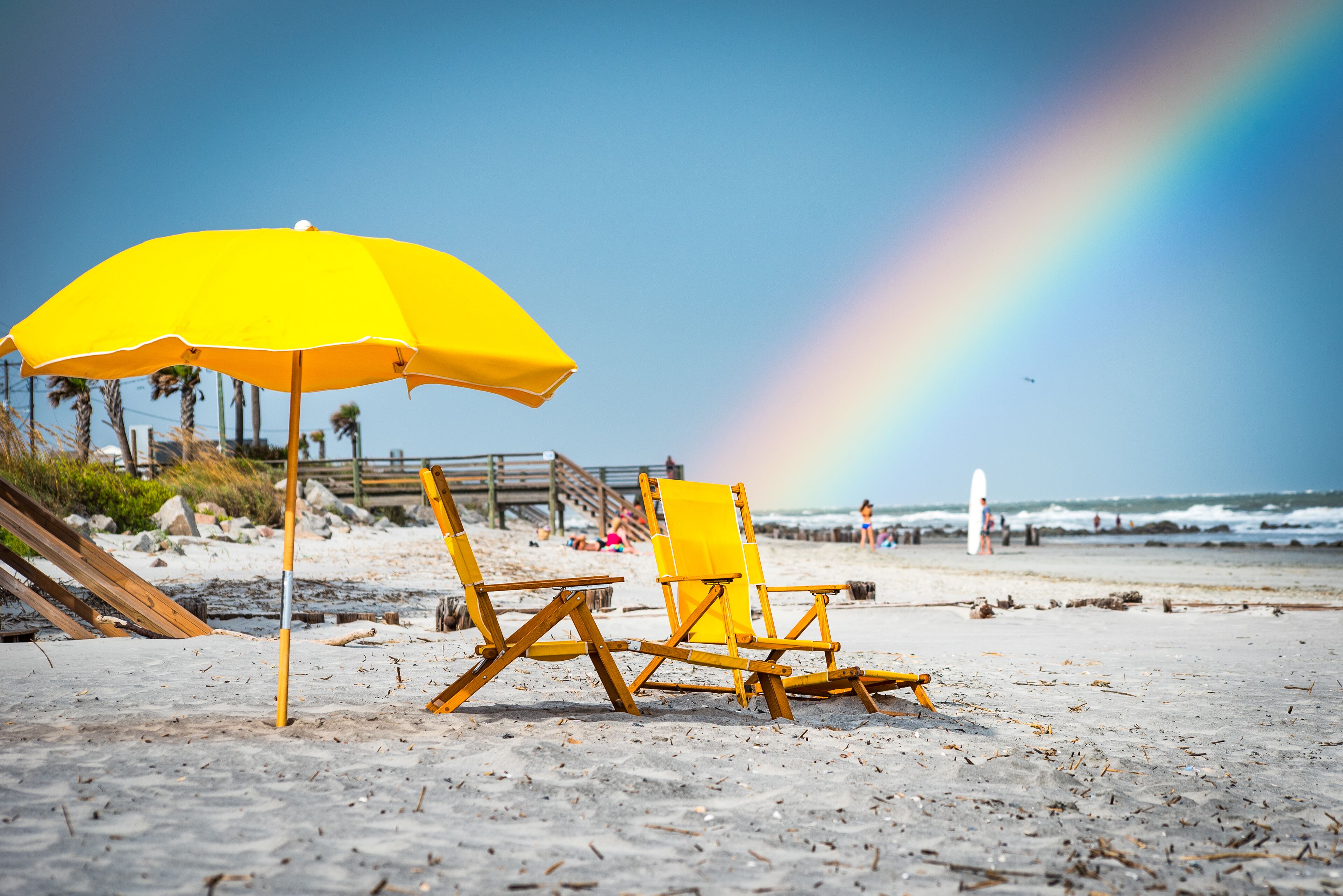 A beach with two wooden beach chairs and a yellow umbrella, a wooden pier, and a rainbow in the background.