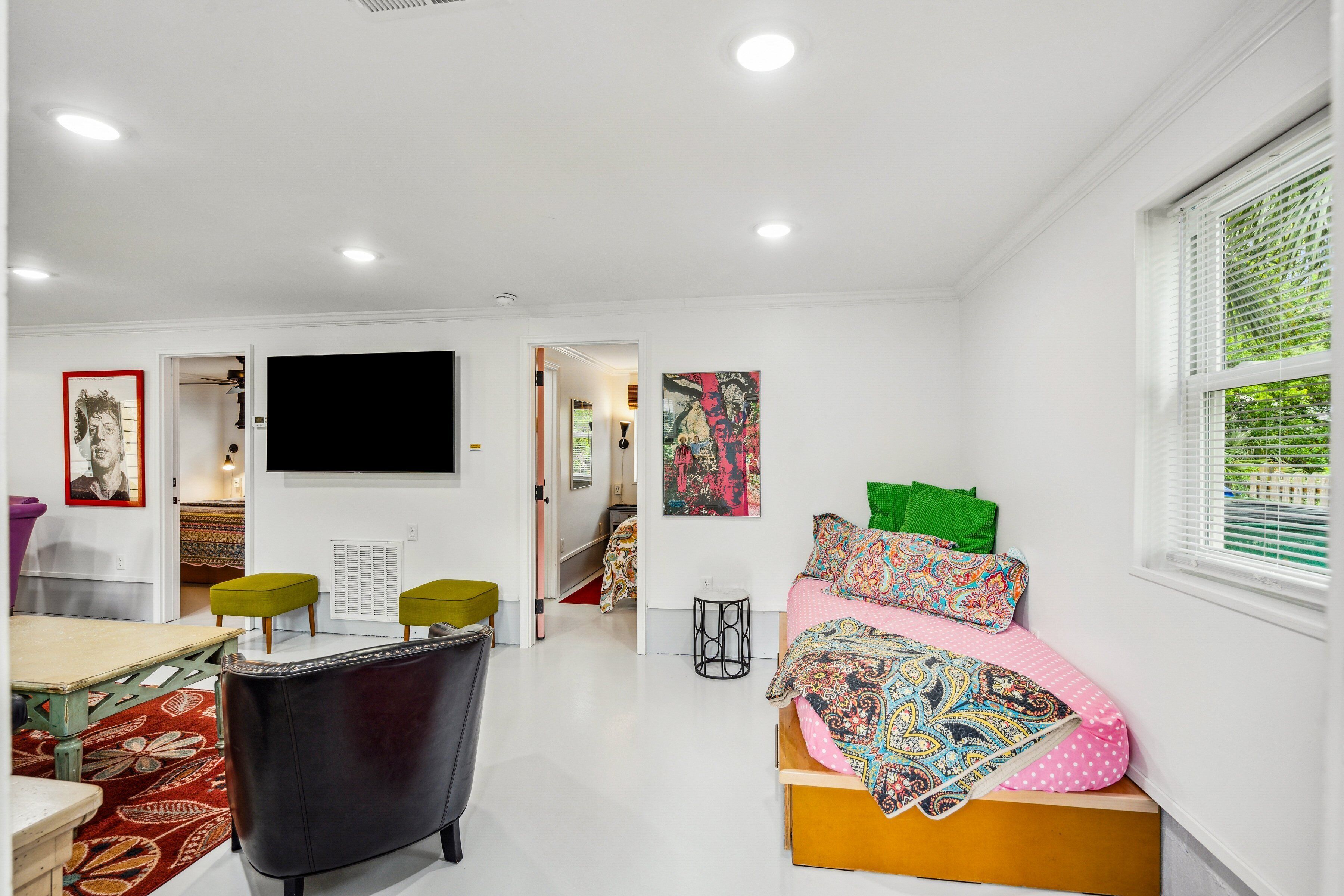 A modern, well-lit room with a bed, a desk, and a chair. There is a window with blinds, a door leading to another room, and a wall-mounted television.