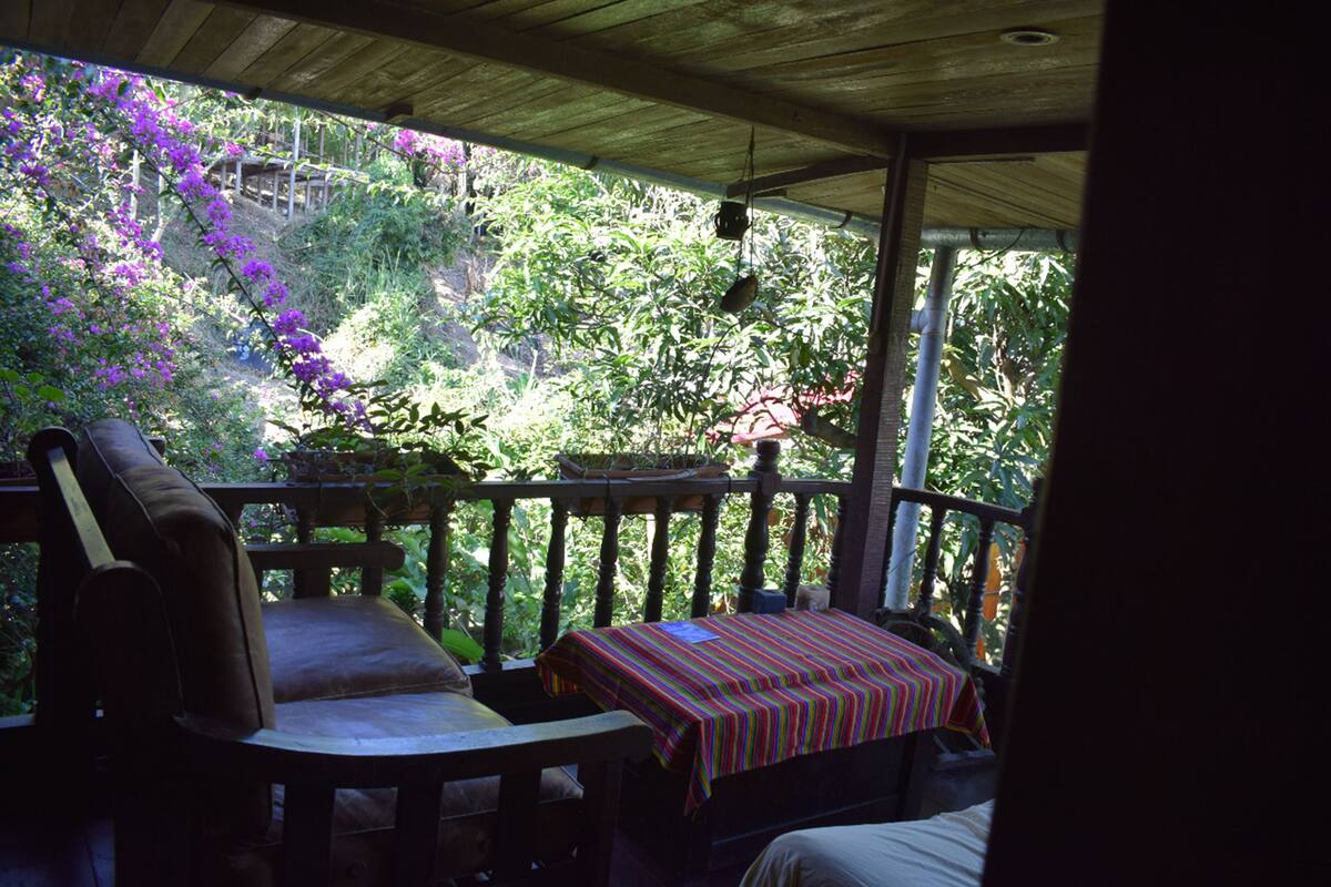 A covered porch with a wooden bench and table, surrounded by lush greenery and purple flowers.