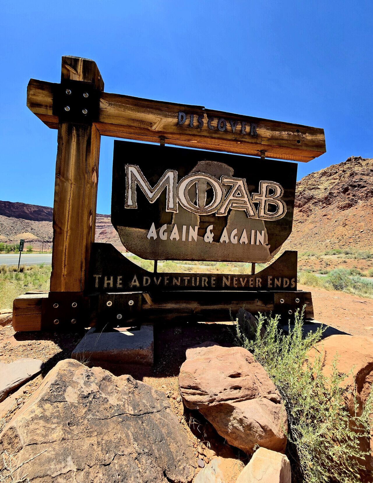 A wooden sign with the text "MOAB AGAIN & AGAIN. THE ADVENTURE NEVER ENDS." in a desert landscape.