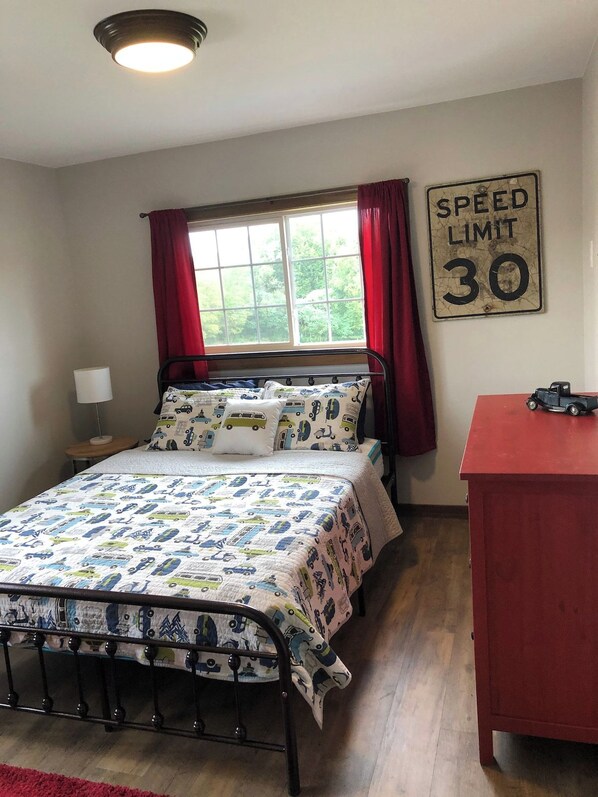 A bedroom with a bed, a window with red curtains, a speed limit sign, and a red dresser. - 1900 FARMHOUSE (Wausau)