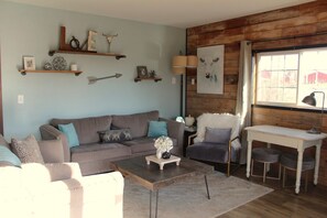 A living room with a sofa, coffee table, and a chair. There are wooden shelves with decorative items and a painting of a cow on the wall.