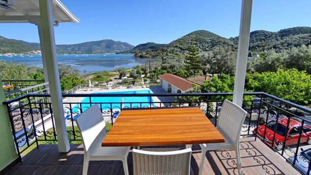 A balcony with a wooden table and chairs, overlooking a swimming pool and a mountainous landscape. - Vliho Bay Suites And Apartments (Lefkada)