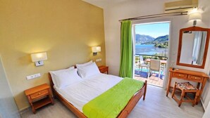 A hotel room with a bed, bedside lamps, a desk, a chair, and a balcony with a view of the sea and mountains. - Vliho Bay Suites And Apartments (Lefkada)