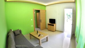 A living room with a green wall, a grey sofa, a wooden coffee table, a television on a wooden stand, and a door leading to another room. - Vliho Bay Suites And Apartments (Lefkada)
