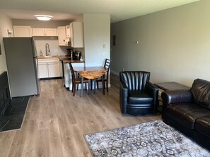 Una cocina moderna con armarios blancos, una mesa de comedor con sillas, un sillón de cuero negro y un sofá de cuero marrón en una sala de estar con alfombra estampada.
