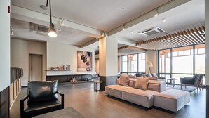 A spacious lobby with a large sectional sofa, a black leather chair, and a small table. There is a large window providing natural light and a view of the outdoors. - Eden Paradise Hotel (Icheon)