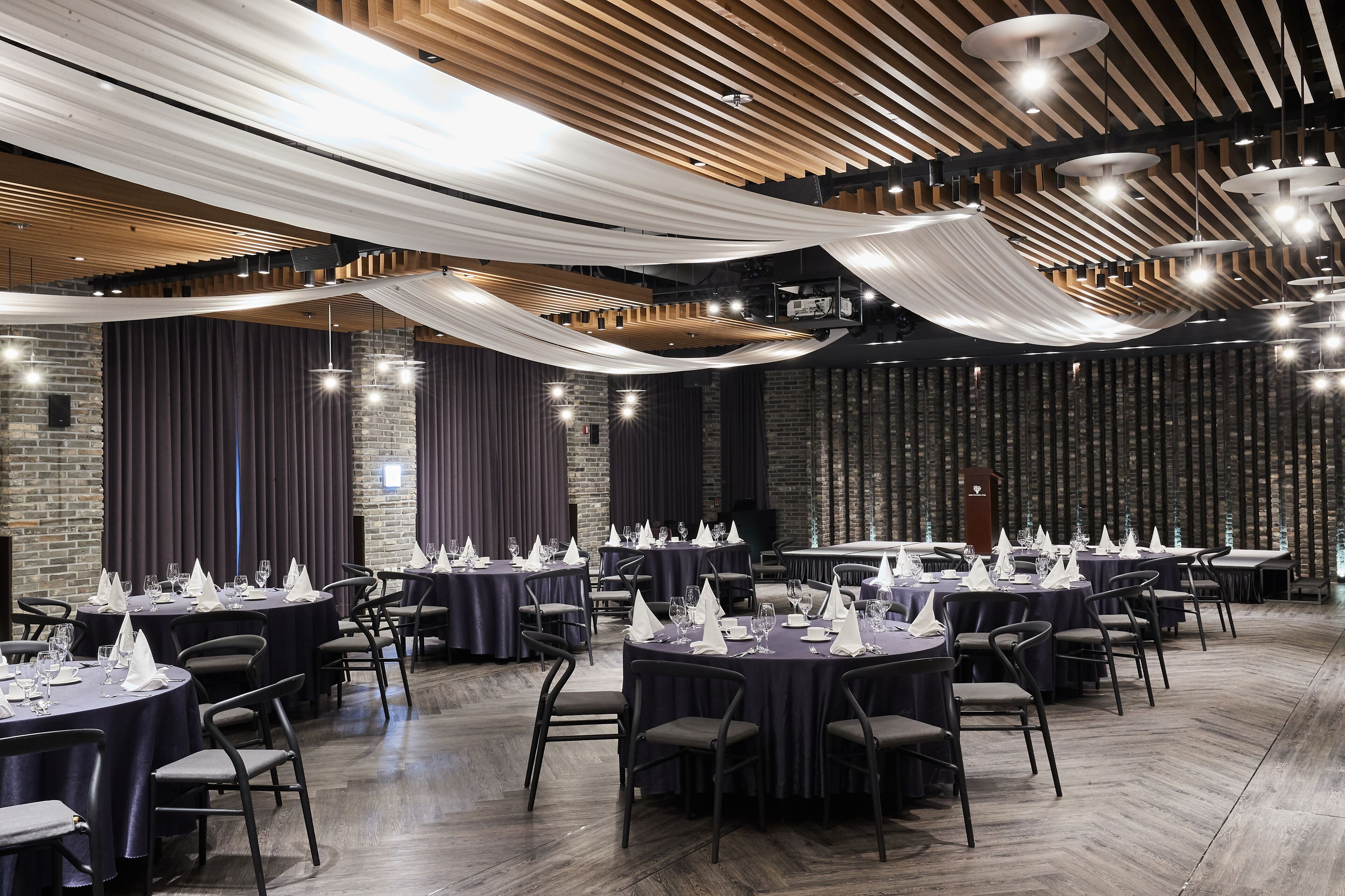 A spacious banquet hall with round tables set for dining, featuring a modern design with wooden beams and hanging fabric accents.