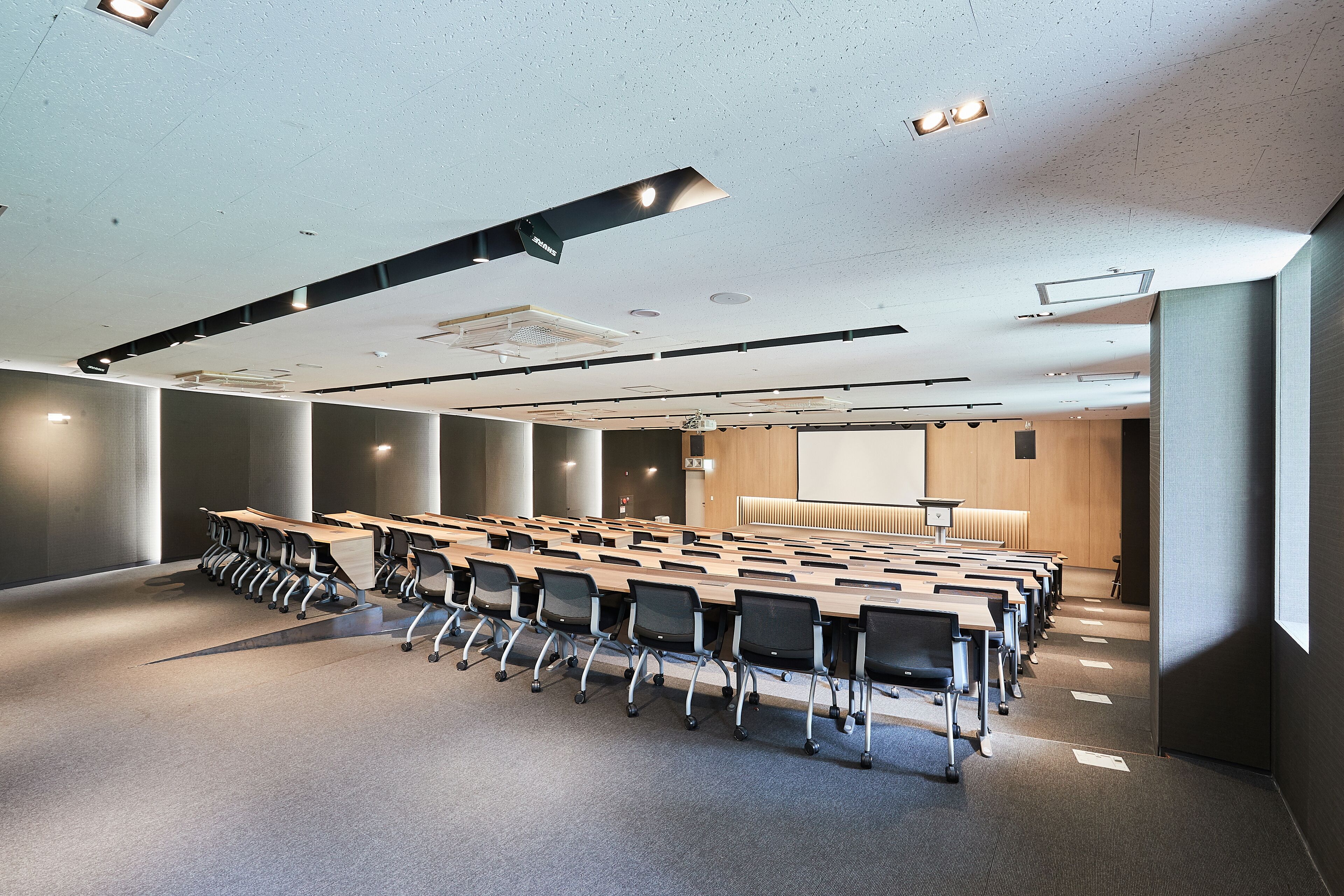 Un salón de conferencias con filas de mesas y sillas, luces en el techo y una pantalla grande.