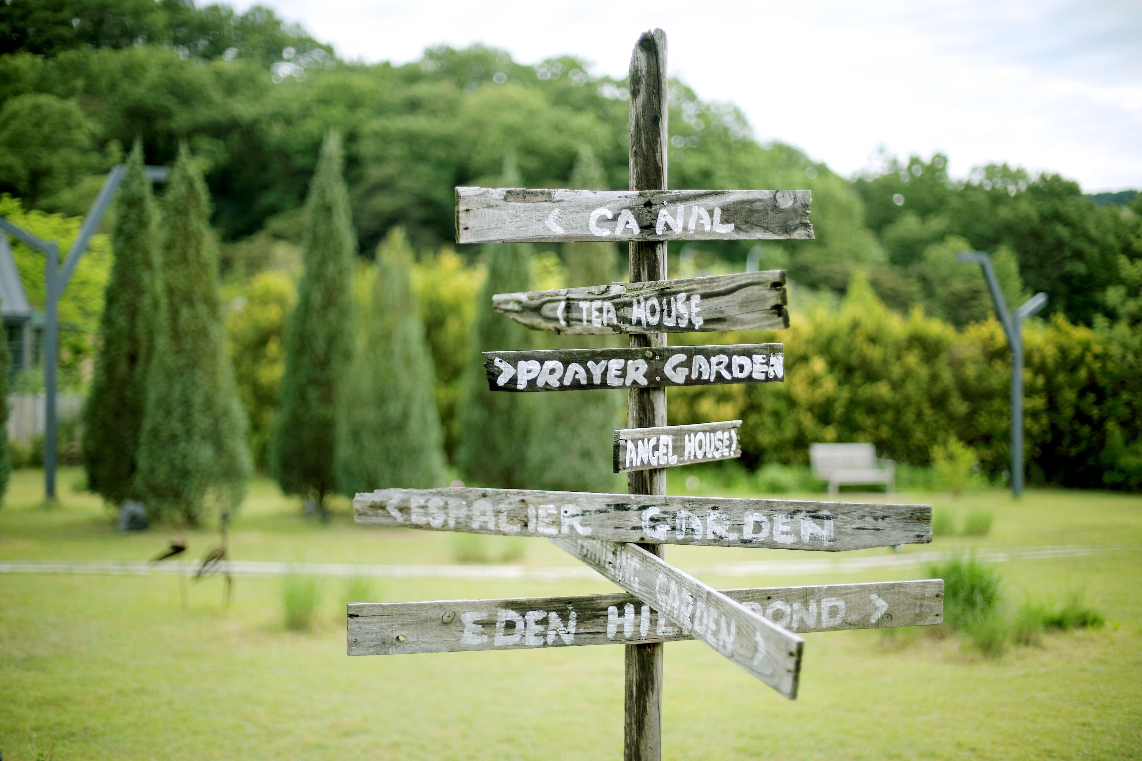 A wooden signpost with multiple directional arrows pointing to various destinations such as "Canal," "Tea House," "Prayer Garden," and "Angel House."