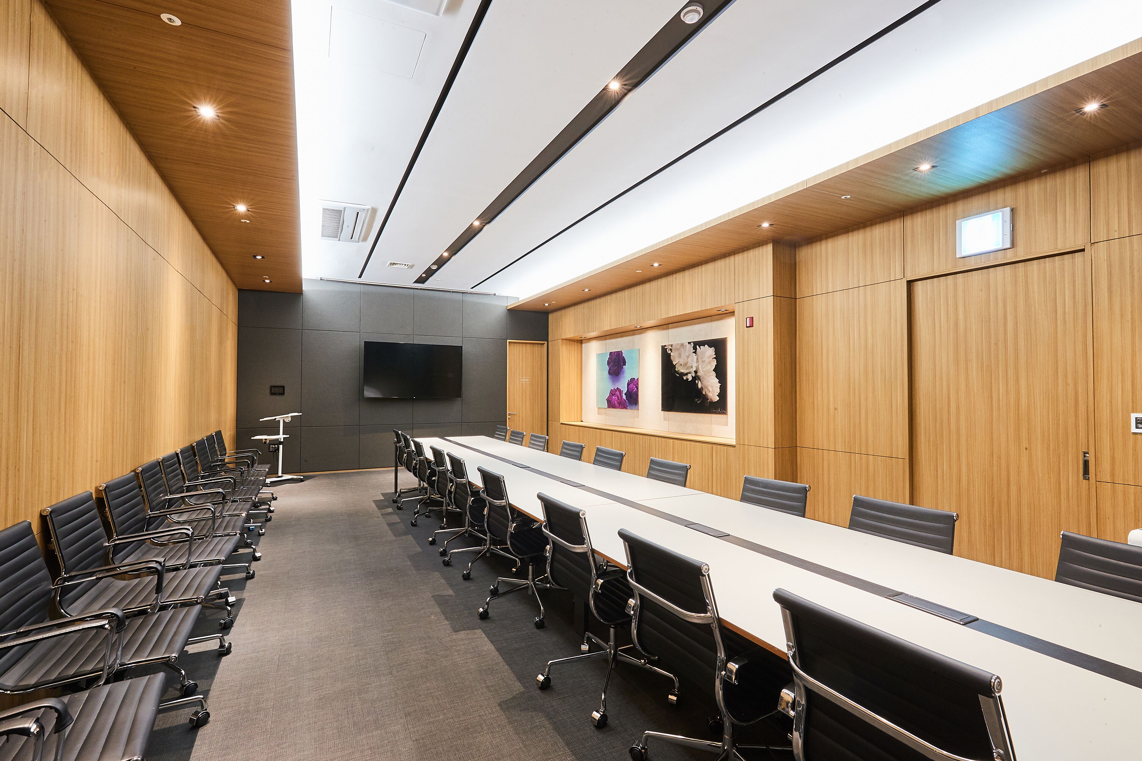 A conference room with a long table, rows of chairs, wooden paneling, and a mounted screen.