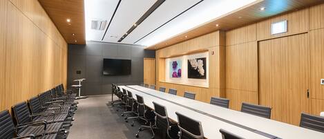 A conference room with a long table, rows of chairs, wooden paneling, and a mounted screen.