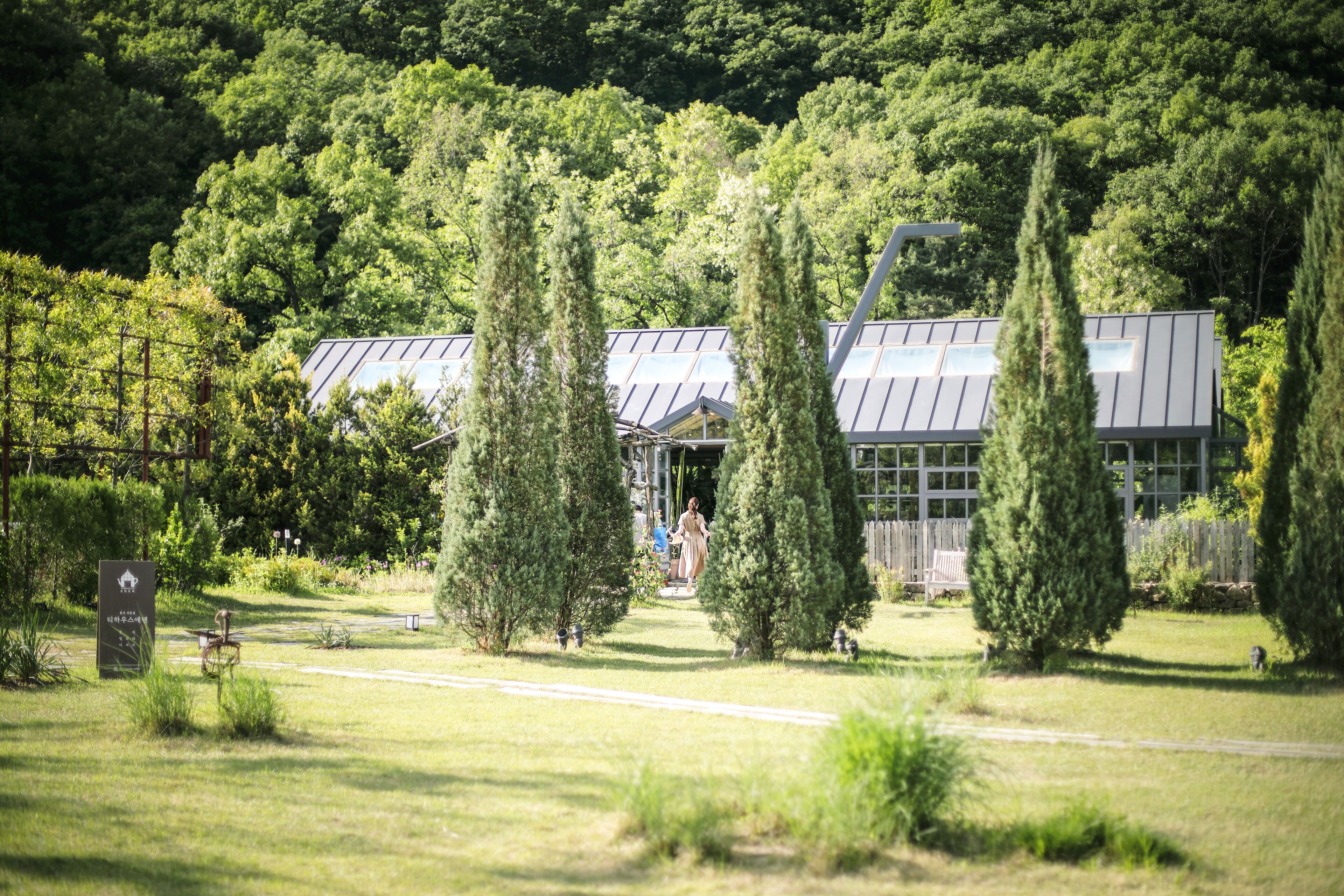 A modern building with a glass facade, surrounded by a well-maintained garden with tall trees and a clear pathway leading to the entrance.