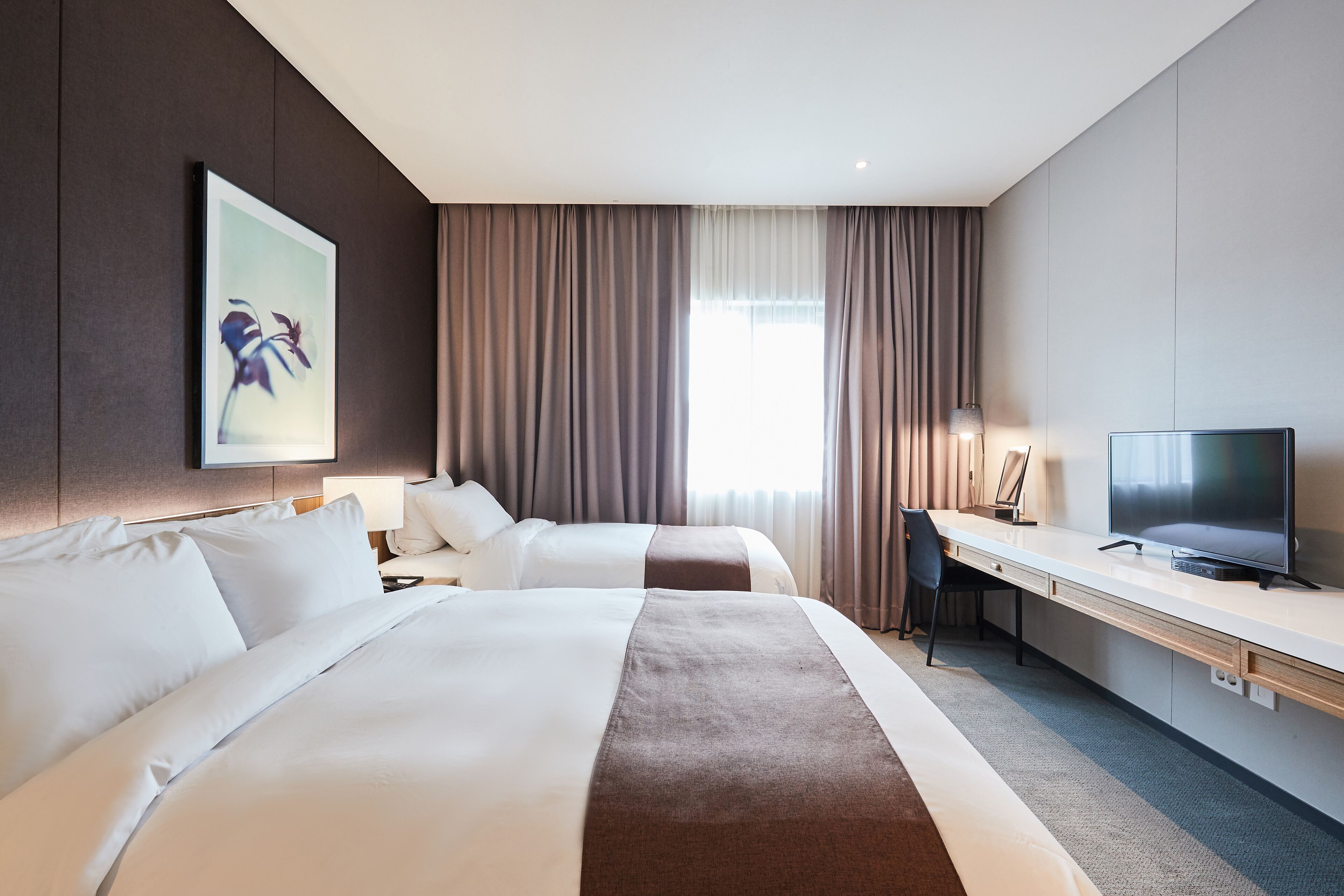 A hotel room with two beds, a desk, a television, and a large window with curtains.