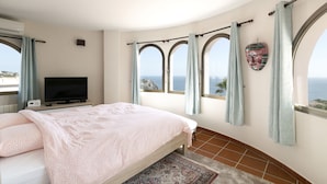 Une chambre à coucher avec un lit, une télévision et une vue sur la mer, visible à travers des fenêtres en arc de cercle.