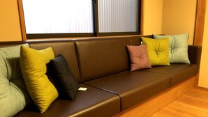 A brown leather sofa with multiple cushions in various colors, positioned in a room with wooden flooring and a window with blinds. - Mokuren (Kyoto)
