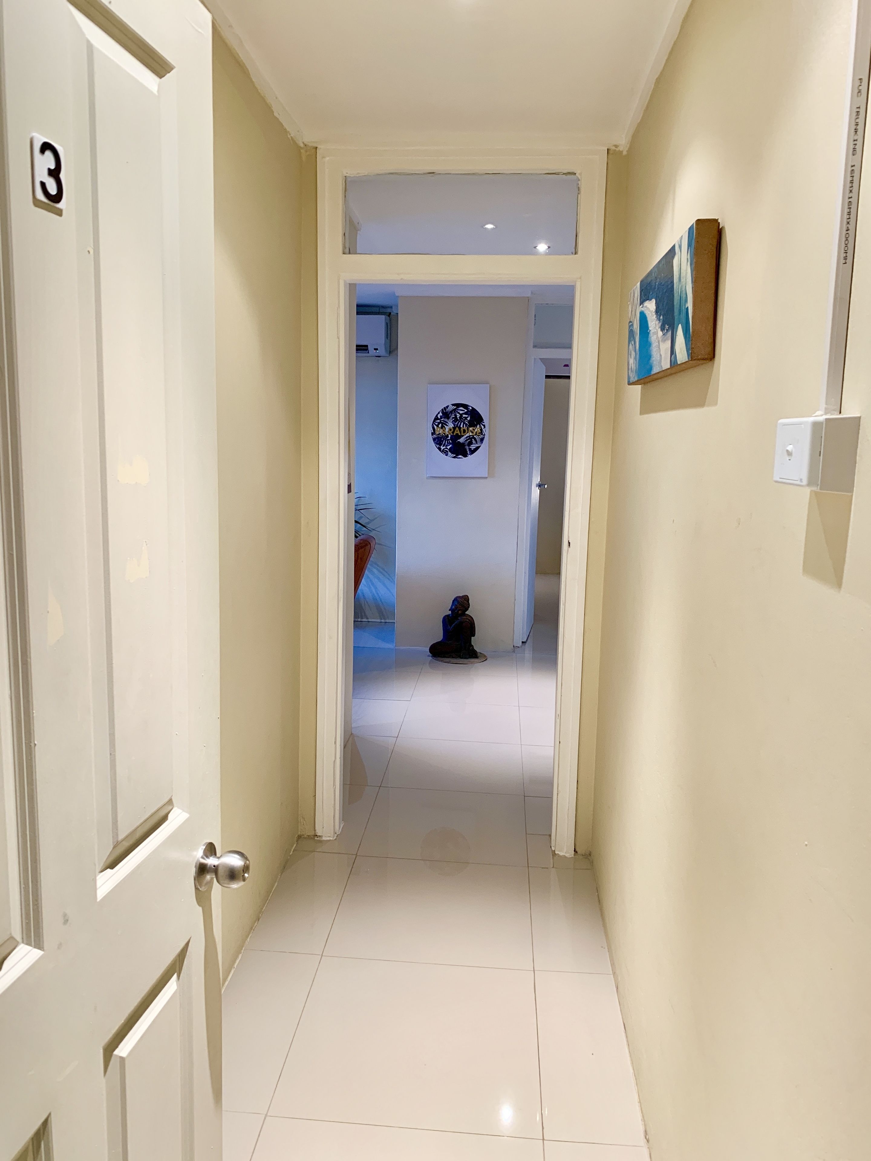 Un corridoio con una porta contrassegnata dal numero '3', un pavimento piastrellato, un interruttore della luce fissato al muro e un quadro incorniciato.