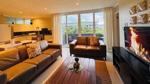 A modern living room with a sofa, a coffee table, and a TV. There is a kitchen in the background with a window overlooking a balcony. - Kidsfirst Apartment 3 (Suva)