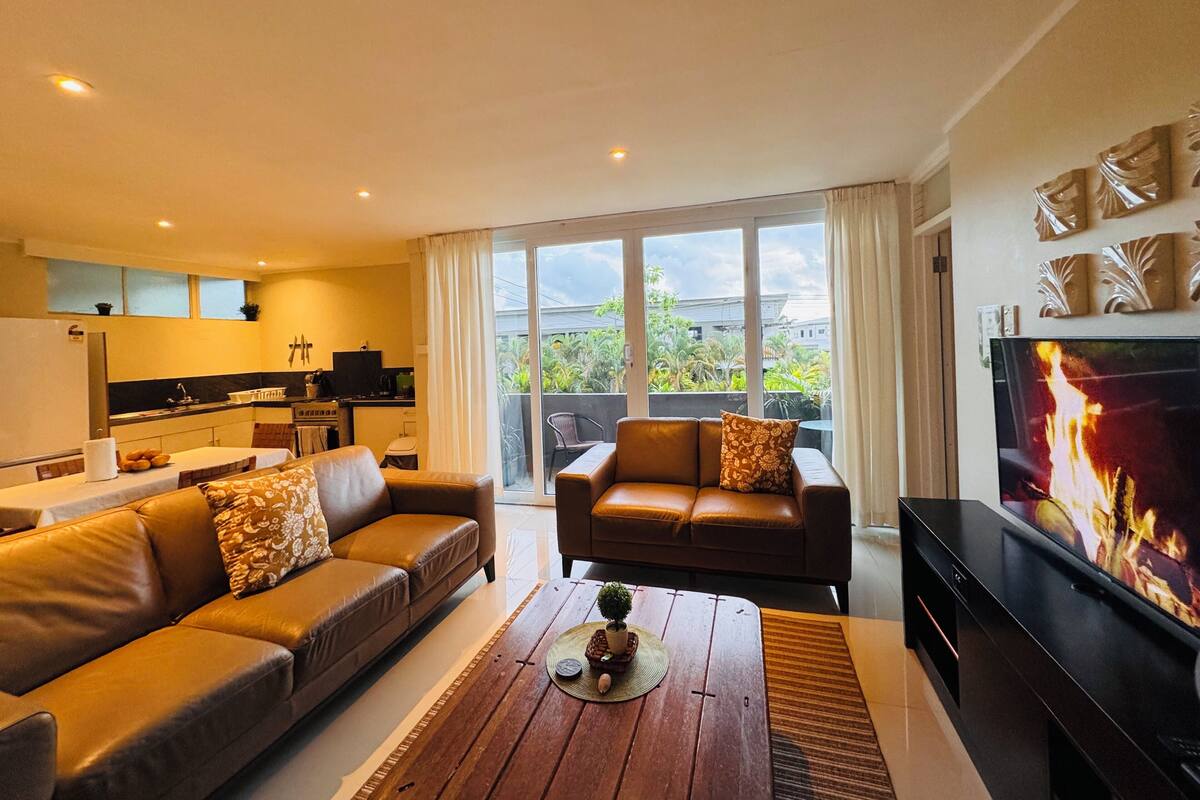 A modern living room with a sofa, a coffee table, and a TV. There is a kitchen in the background with a window overlooking a balcony.