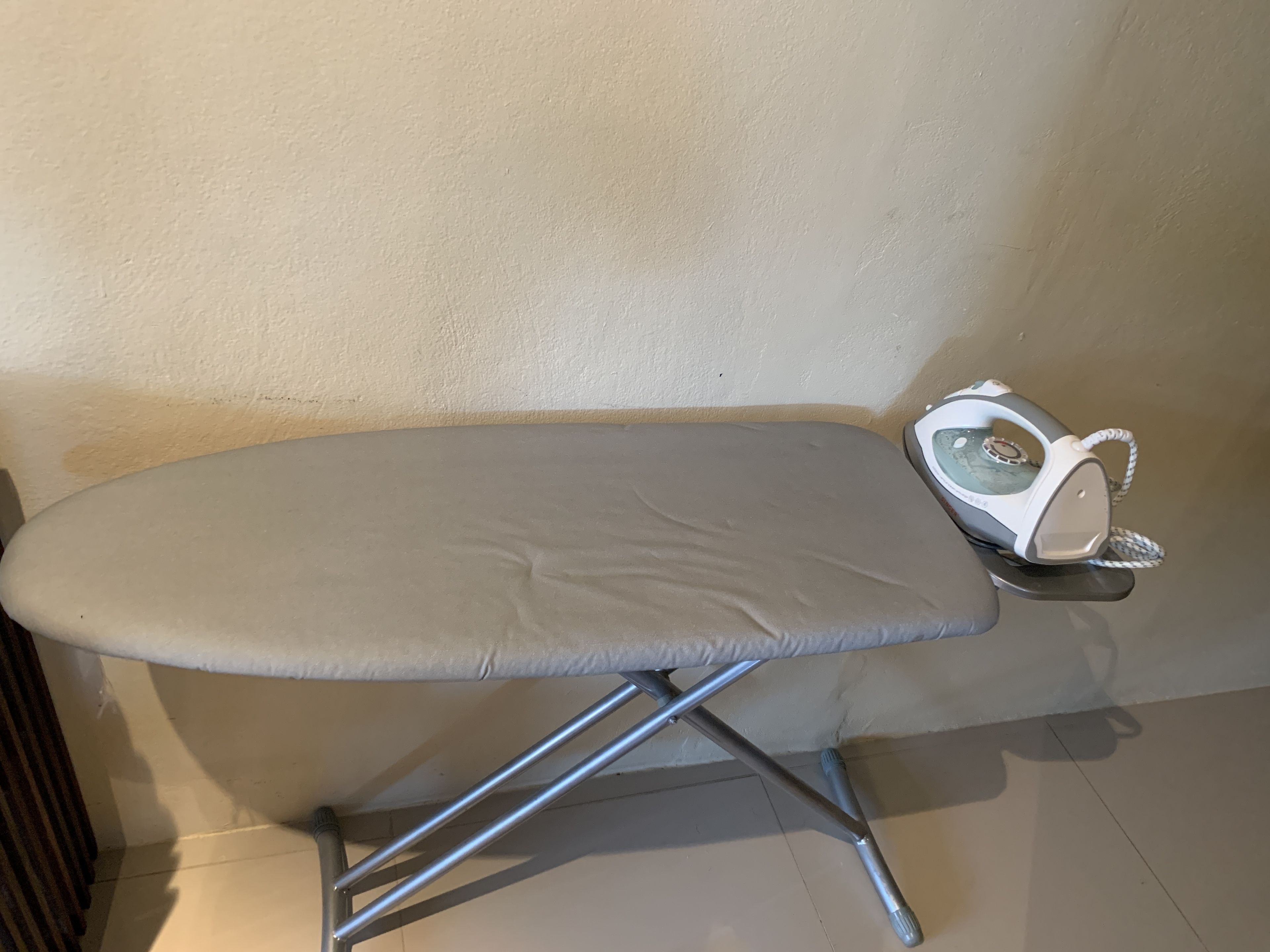 An ironing board with a grey pad and a white iron in a corner of a room with beige walls and a tiled floor.