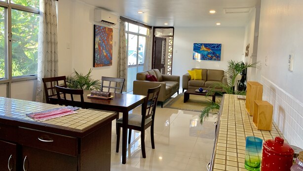 Un espacio habitable amplio con una mesa de comedor, sillas, un sofá y una mesa de centro. Hay dos cuadros en las paredes, una ventana con cortinas y una puerta que da a otra habitación.