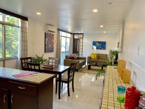 Un espacio habitable amplio con una mesa de comedor, sillas, un sofá y una mesa de centro. Hay dos cuadros en las paredes, una ventana con cortinas y una puerta que da a otra habitación.