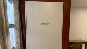 Una habitación con pared de paneles de madera, una puerta blanca con manija, una ventana con cortinas estampadas y una mesa con sillas.