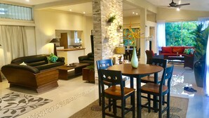 A modern living room with a sofa, coffee table, and dining table with chairs. There is a kitchen in the background and a red sofa in the dining area. - The Williamson House (Suva)