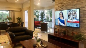 A modern living room with a television displaying a news program, a comfortable sofa, a coffee table with remote controls, and a view of a balcony with plants. - The Williamson House (Suva)