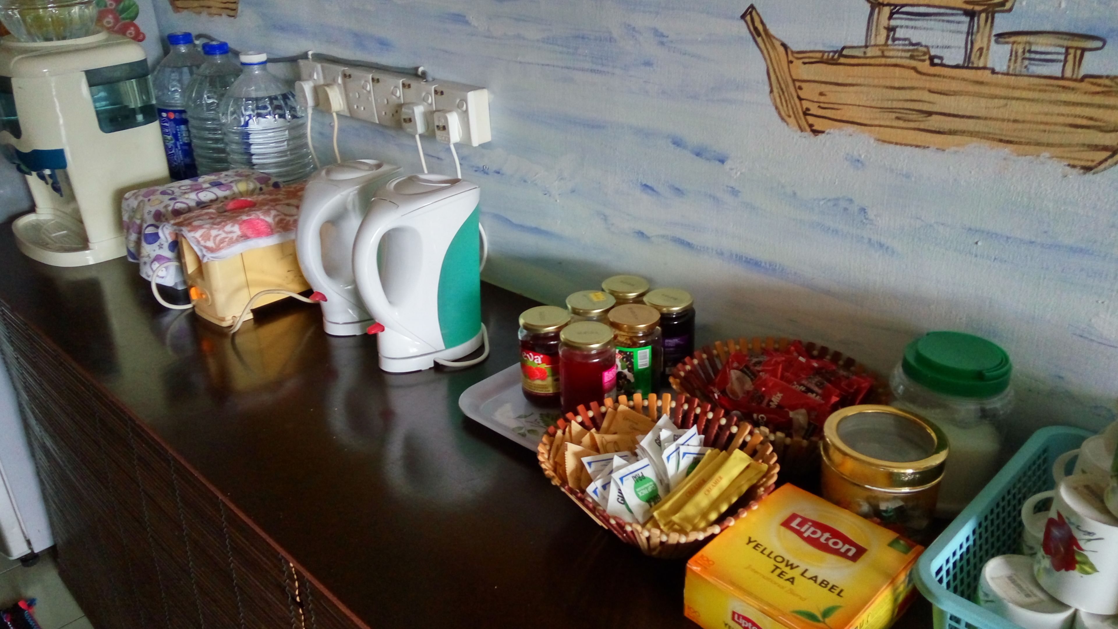 A kitchen counter with a coffee maker, a kettle, jars of jam, a basket of cigarettes, a box of Lipton tea, and a green basket with cups.