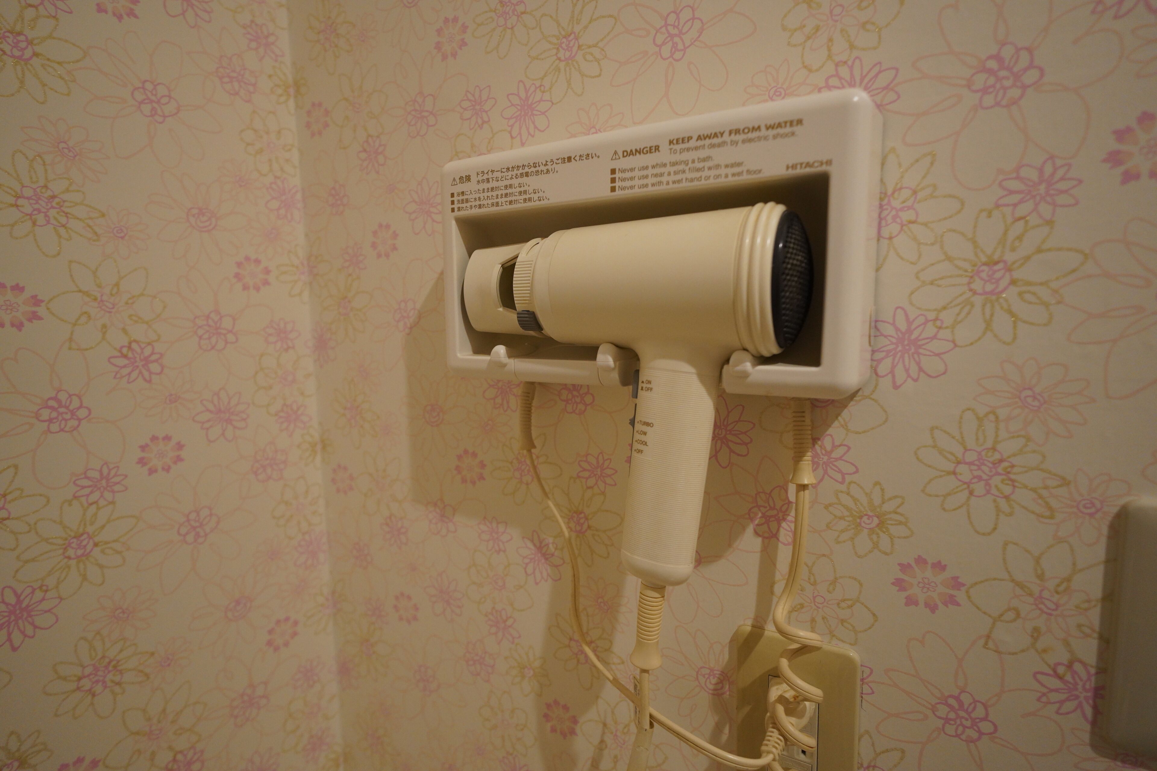 A hair dryer mounted on a wall with a floral wallpaper pattern.