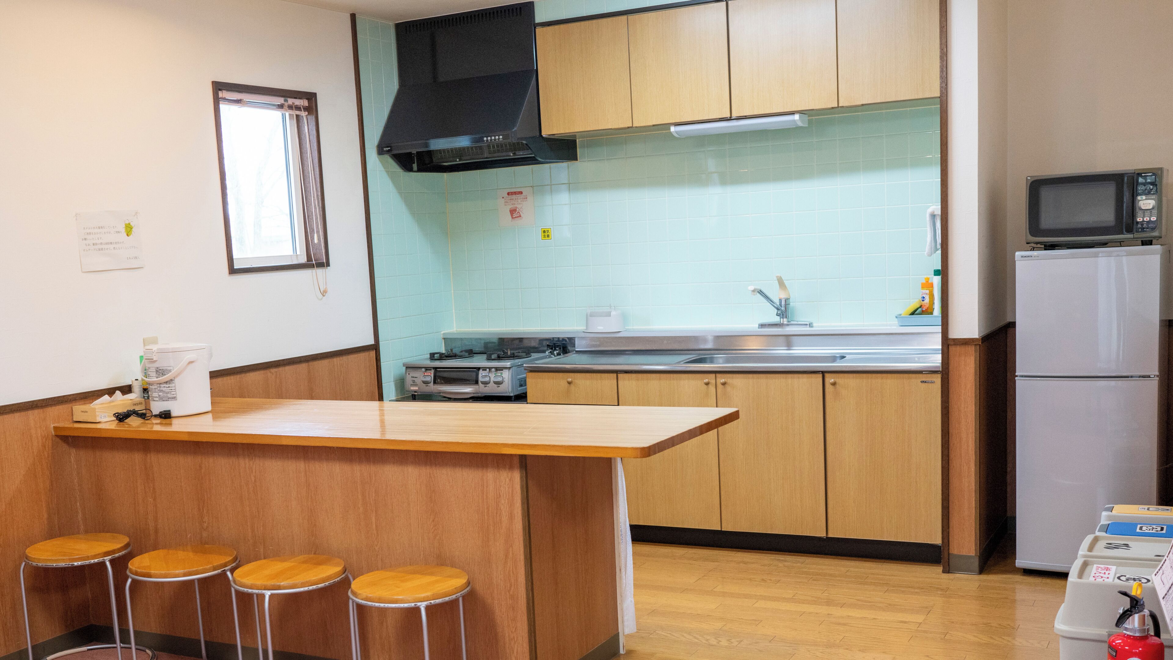 Una cocina con armarios de madera, un lavamanos de acero inoxidable, un microondas y un refrigerador. Hay taburetes en la barra y una ventana sobre el lavamanos.