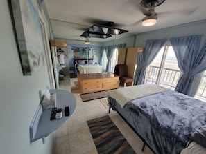 Quarto com cama, cómoda de madeira, escrivaninha com cadeira, ventoinha no teto e vista para o oceano pela janela.