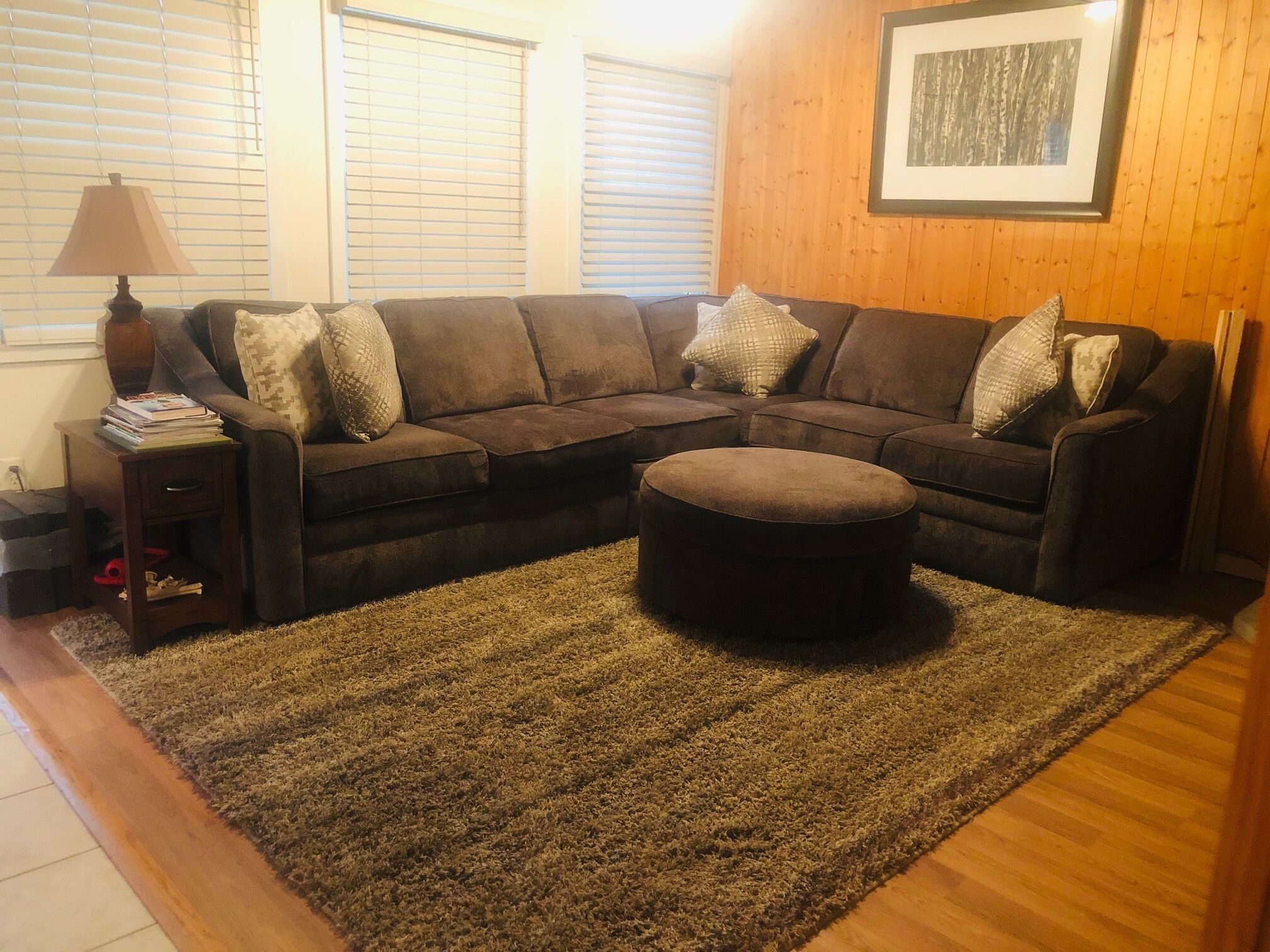 A living room with a sectional sofa, a round ottoman, a small side table, and a lamp.